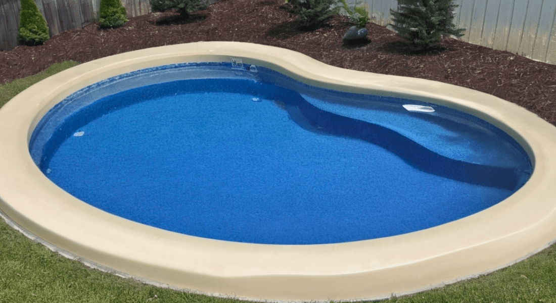 An oval-shaped swimming pool with clean blue water, surrounded by a beige edge, sits in a landscaped yard with some small trees and a fence in the background.