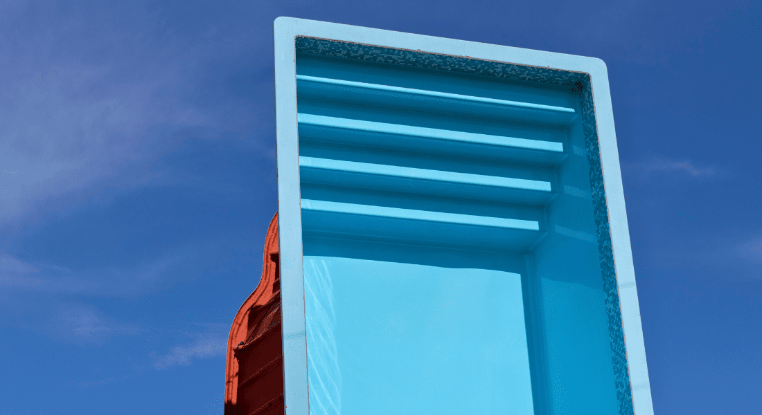 A large, empty, blue rectangular swimming pool structure is shown standing on its side against a clear blue sky. Fiberglass pool shell
