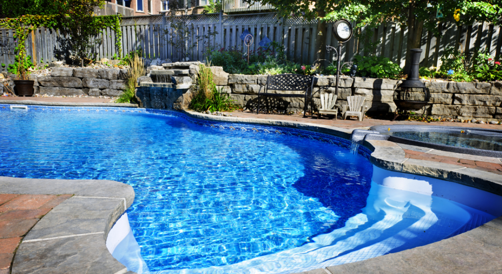 A backyard pool with clear blue water, adjacent to a stone retaining wall, bench, and greenery. A hot tub is visible on the right, and a fence lines the background.