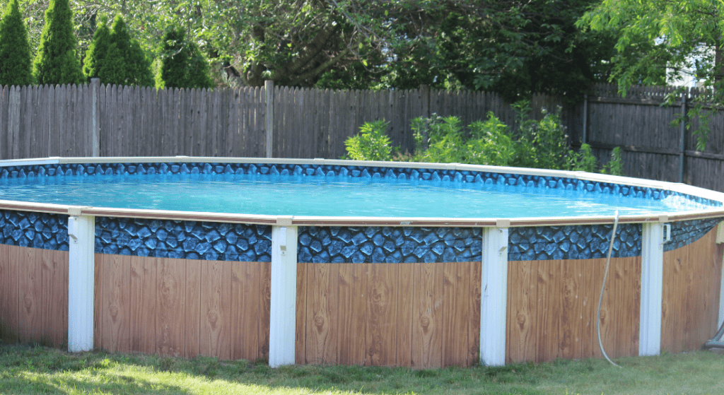 Above-ground swimming pool with blue water and a patterned exterior in a backyard, surrounded by a wooden fence and green foliage.