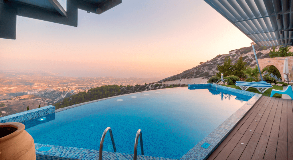 Infinity pool on a wooden deck with a view of a valley and distant mountains at sunset. Lounge chairs and greenery are nearby.