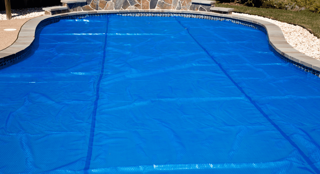 A swimming pool covered with a blue solar pool cover, bordered by stone and tile. pool trend 2024