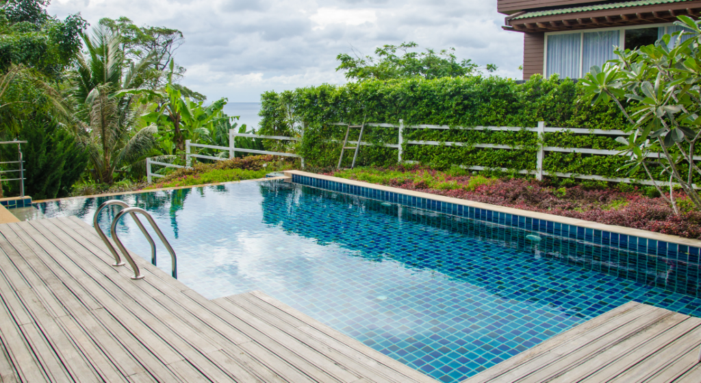 A rectangular outdoor pool with blue tiles is surrounded by a wooden deck. Lush greenery and a partial view of the ocean are in the background. Your Dream Pool & Spa with Sutton Pools in Sango, TN