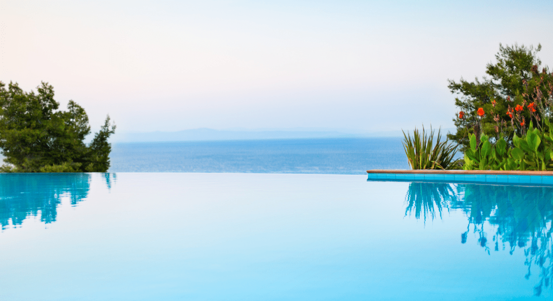 Infinity pool overlooking the ocean, surrounded by trees and flowering plants under a clear sky. pool trend 2024