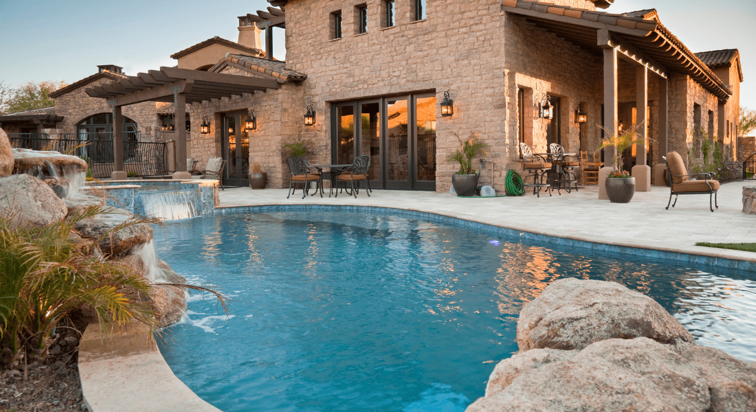 A luxurious stone house with outdoor seating, a tiled patio, and a swimming pool featuring a waterfall. pool trend 2024