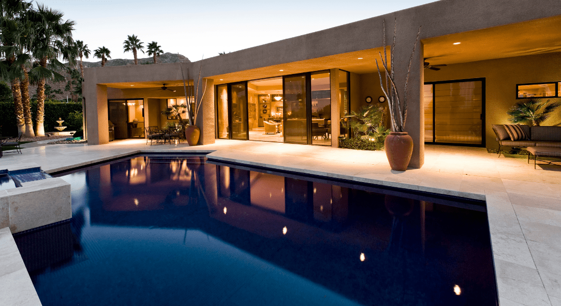 Modern house with large glass doors opening to a patio and a rectangular pool, surrounded by potted plants and outdoor furniture, set against a backdrop of palm trees at dusk. pool trend 2024