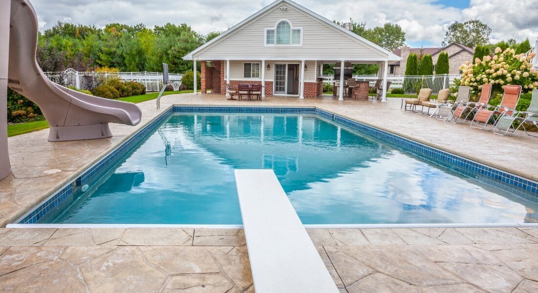 A backyard pool with a slide and diving board, surrounded by lounge chairs. A house with a patio is in the background, set against a backdrop of trees and a cloudy sky.