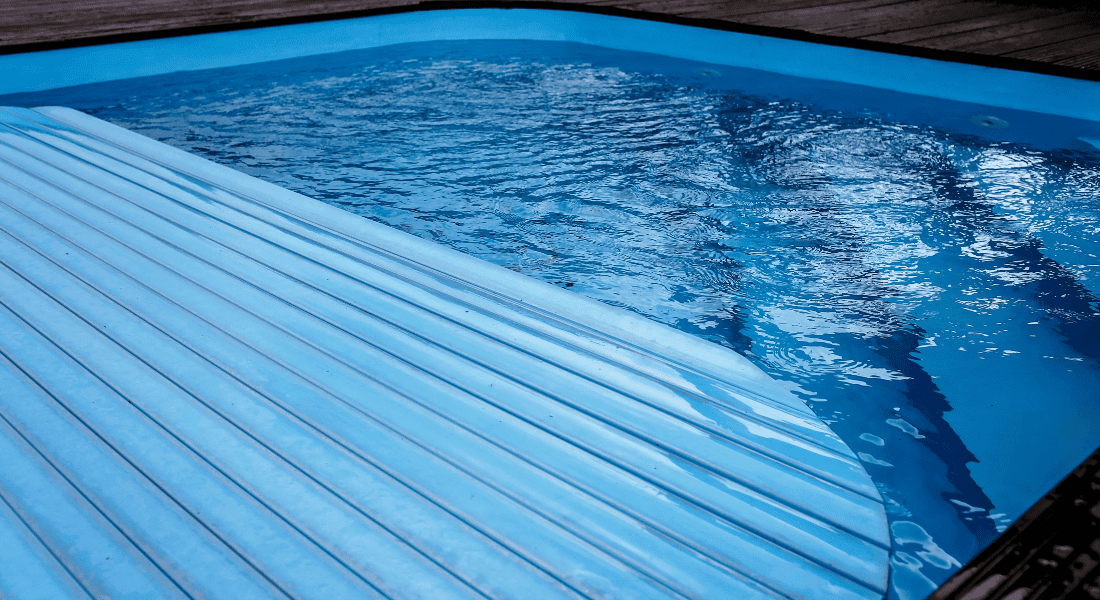 A swimming pool with a partially closed white slatted safety cover. The pool water is clear and the edge has a wooden deck.