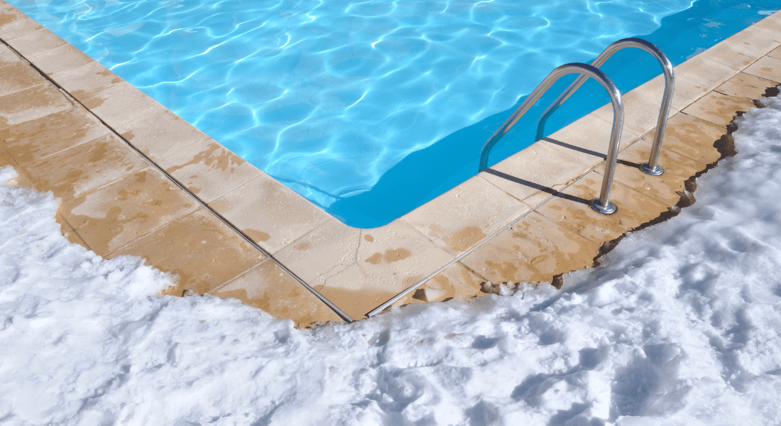 A swimming pool with clear blue water is adjacent to a snowy surface. The pool ladder is visible on the right.