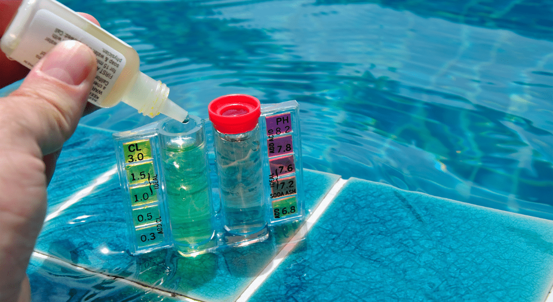 A hand is adding drops to a chlorine and pH test kit beside a swimming pool, with water visible in the background.