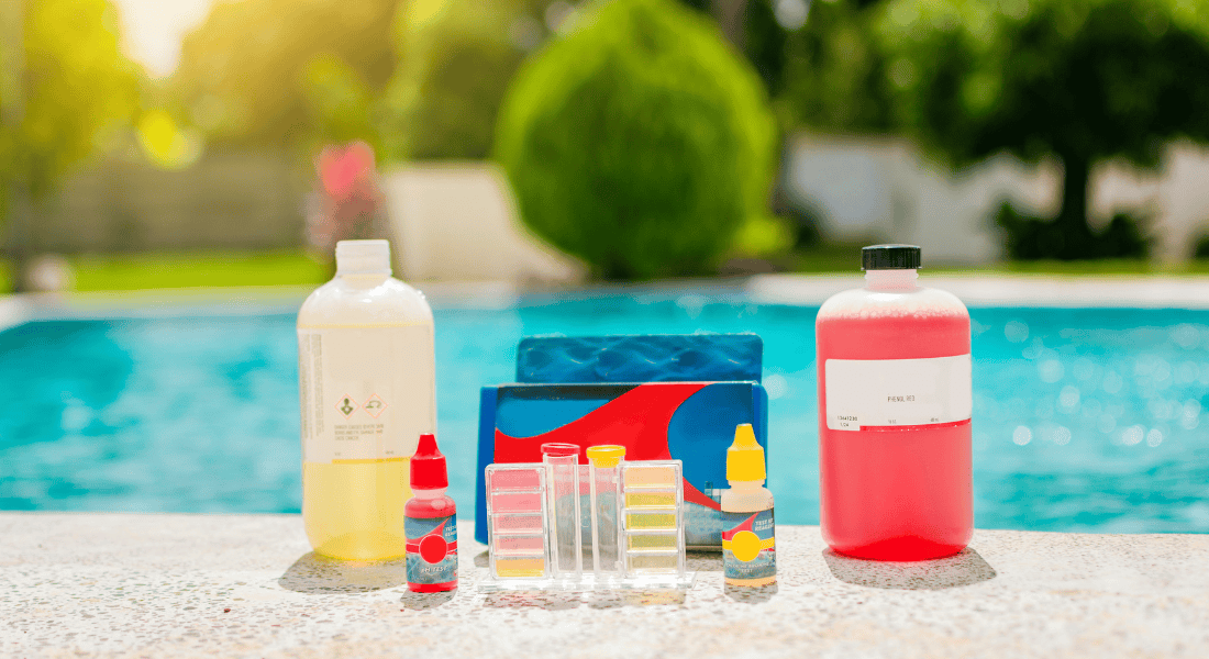 Various pool maintenance products, including two large bottles, test kits, and smaller bottles, are displayed on a surface near a swimming pool.