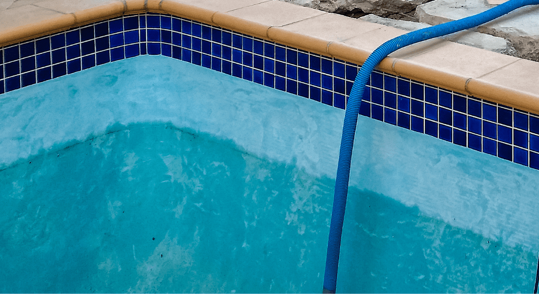 Empty swimming pool with blue hose inside, blue mosaic tiles along the upper edge, and a concrete deck surrounding it.