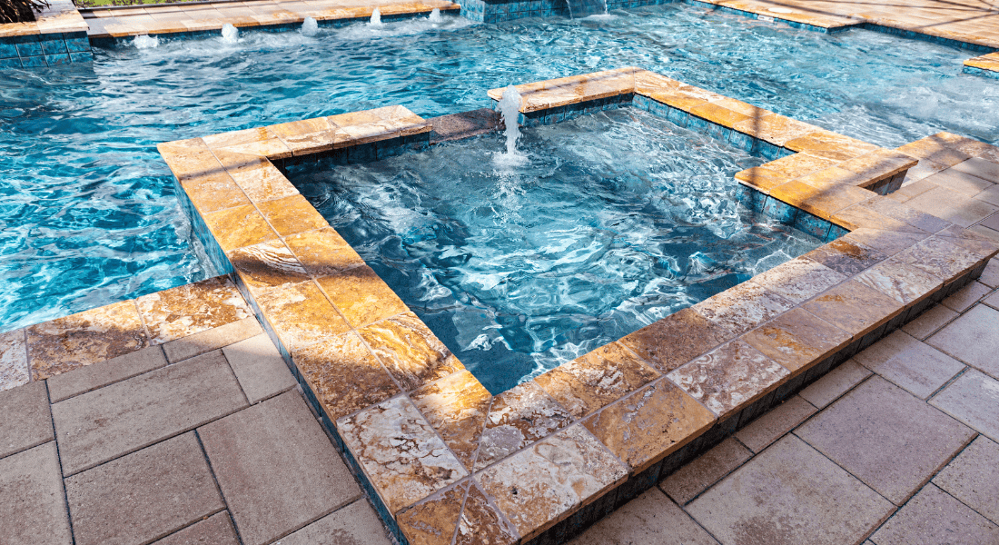 A tiled spa with a small fountain sits adjacent to a swimming pool, both surrounded by tan stone pavers.